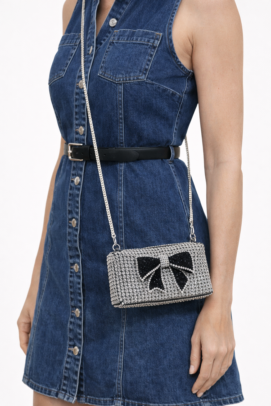 Woman wearing a denim dress with a black belt, holding a decorative handbag with a bow design.